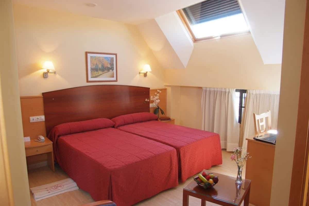 Quarto de hotel com uma cama grande, duas mesas de cabeceira, uma escrivaninha, uma cadeira, uma pequena mesa com frutas, um vaso com flores e um quadro na parede.