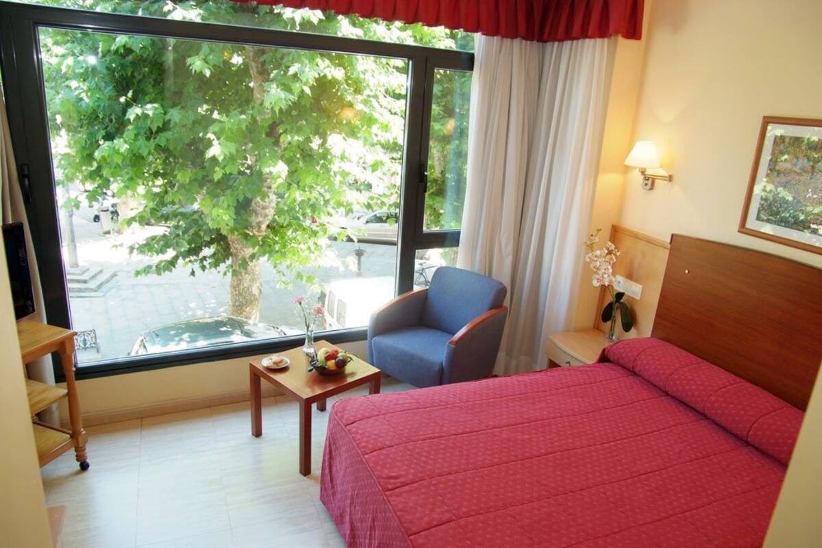 Quarto de hotel com uma janela grande, cama, cadeira, pequena mesa com um vaso de flores e mesa de cabeceira com abajur.