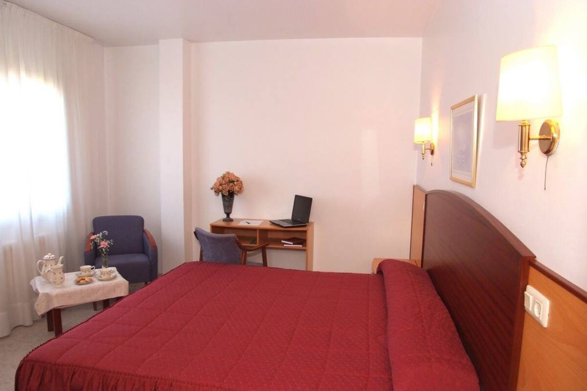 Quarto de hotel com uma cama grande, uma escrivaninha com um laptop, uma cadeira, uma mesinha de centro com chá e um vaso com flores.