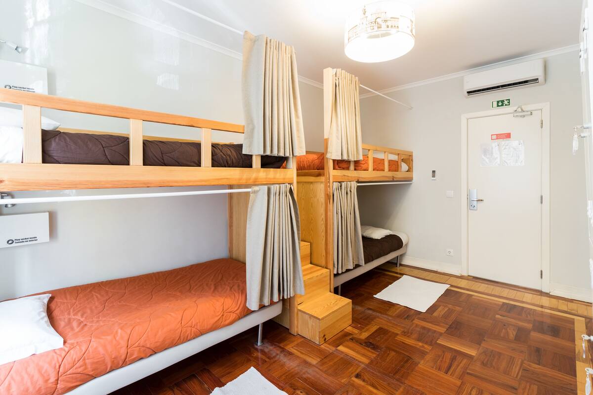 Een slaapzaal met stapelbedden, een houten vloer, een witte deur met een nooduitgangsbordje, en een plafondlamp.