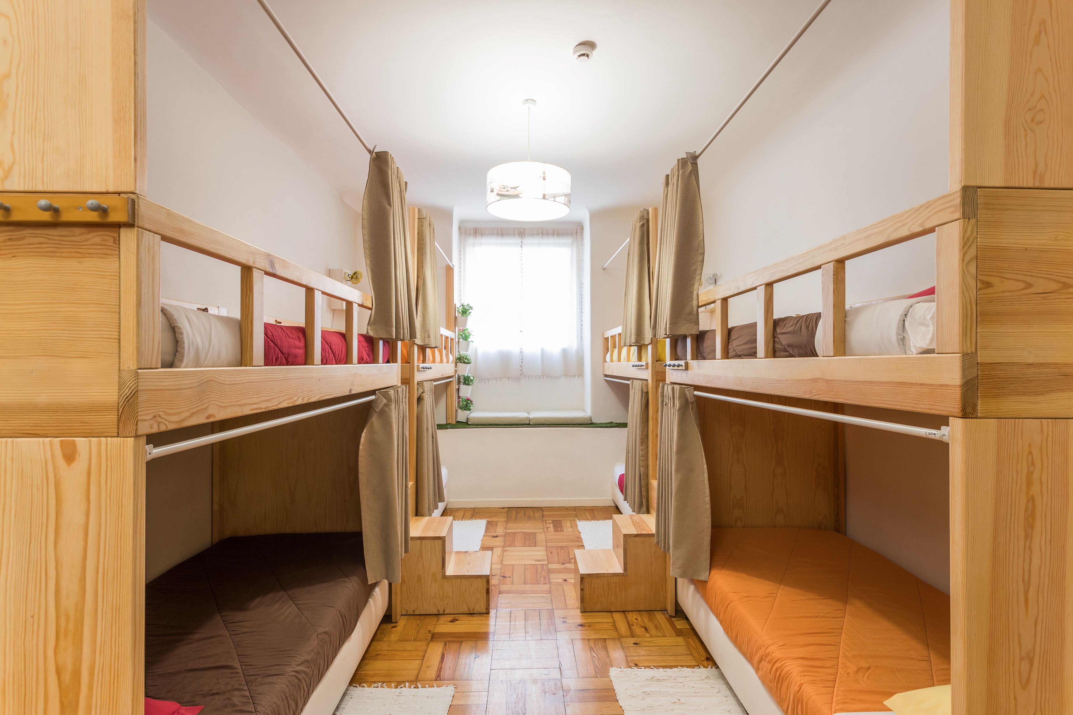 A narrow room with two bunk beds, wooden walls, a window with curtains, and a ceiling light.