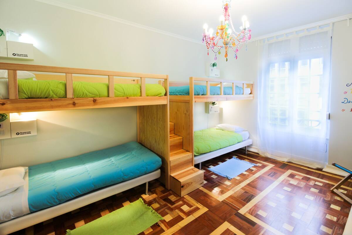 Een slaapzaal met stapelbedden, een houten trap, een kroonluchter en een raam met gordijnen.