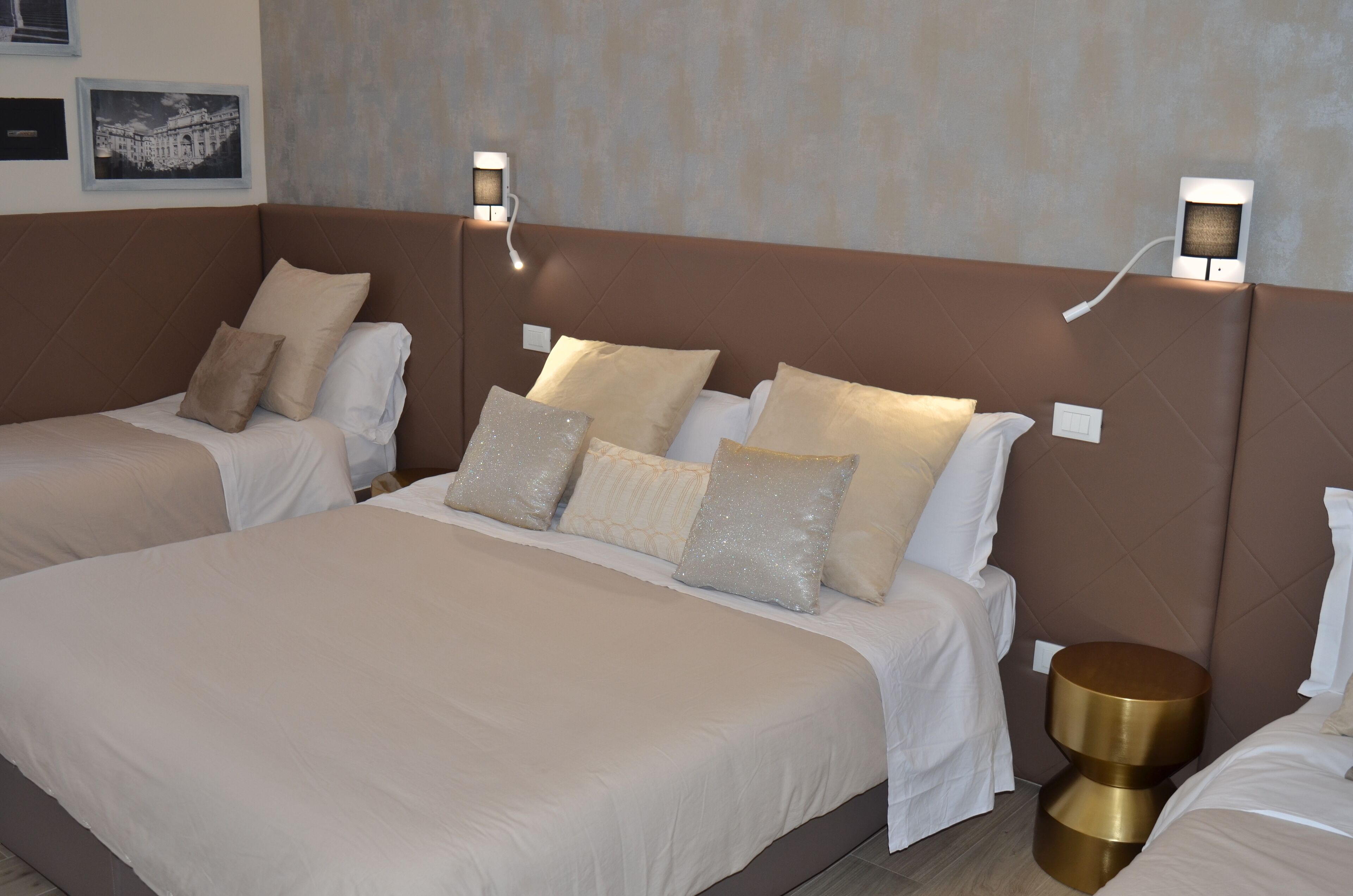 Hotelkamer met twee bedden, beige beddengoed, decoratieve kussens, wandlampen en een klein rond tafeltje.