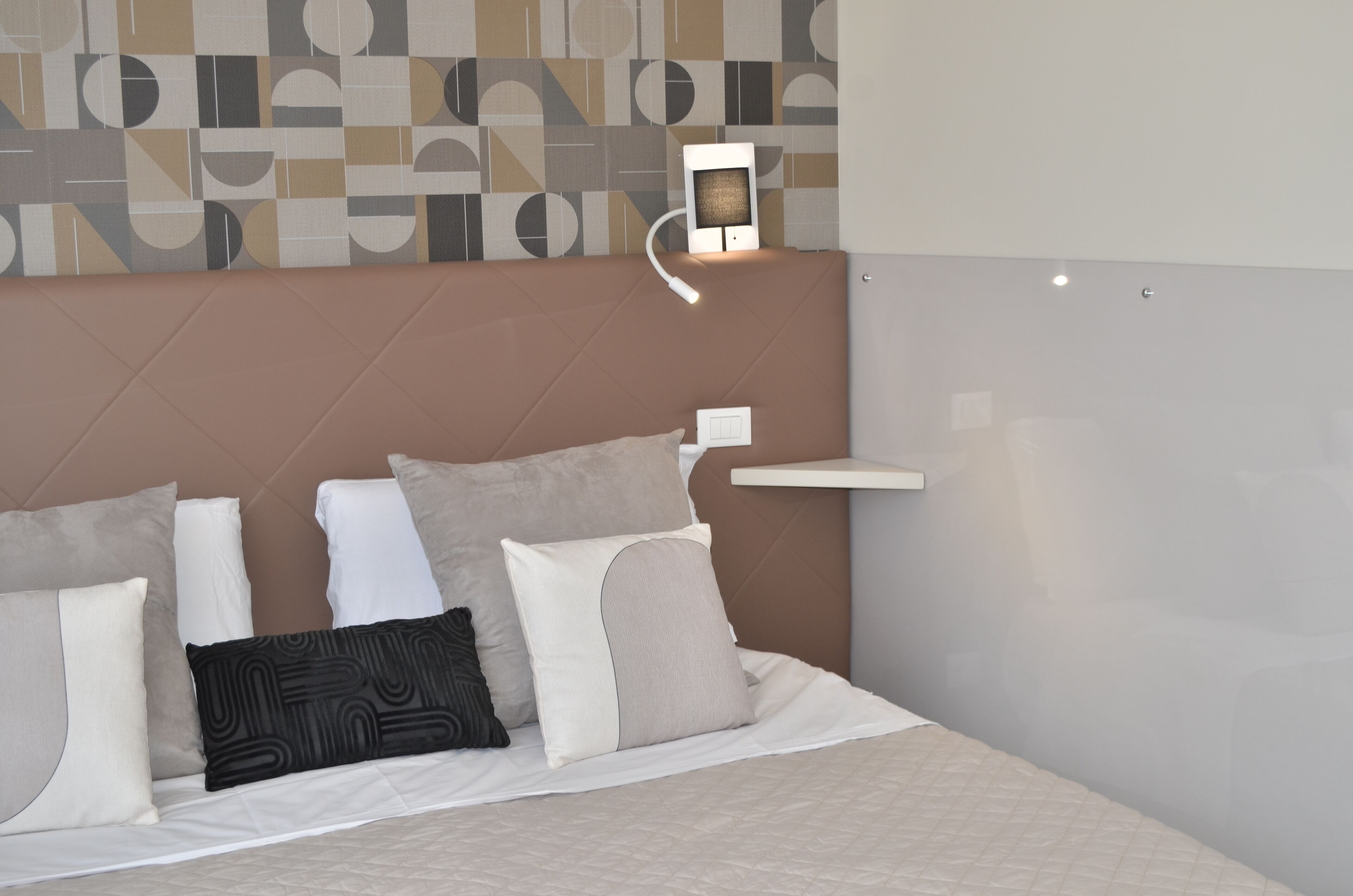 Et moderne hotelværelse med en stor seng, flere puder, en sengegavl med geometrisk mønster, et natbord og en vægmonteret lampe.