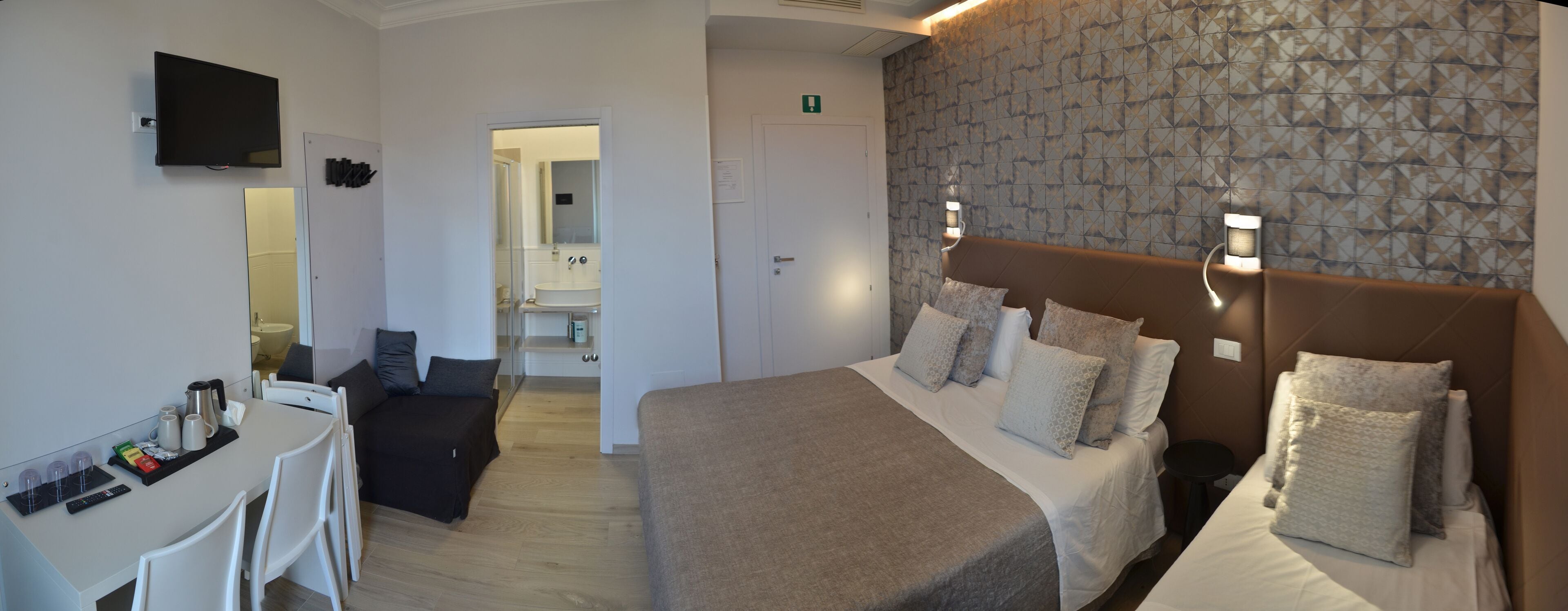 A hotel room with a bed, bedside tables, a desk with a chair, a television, and a bathroom visible through an open door.