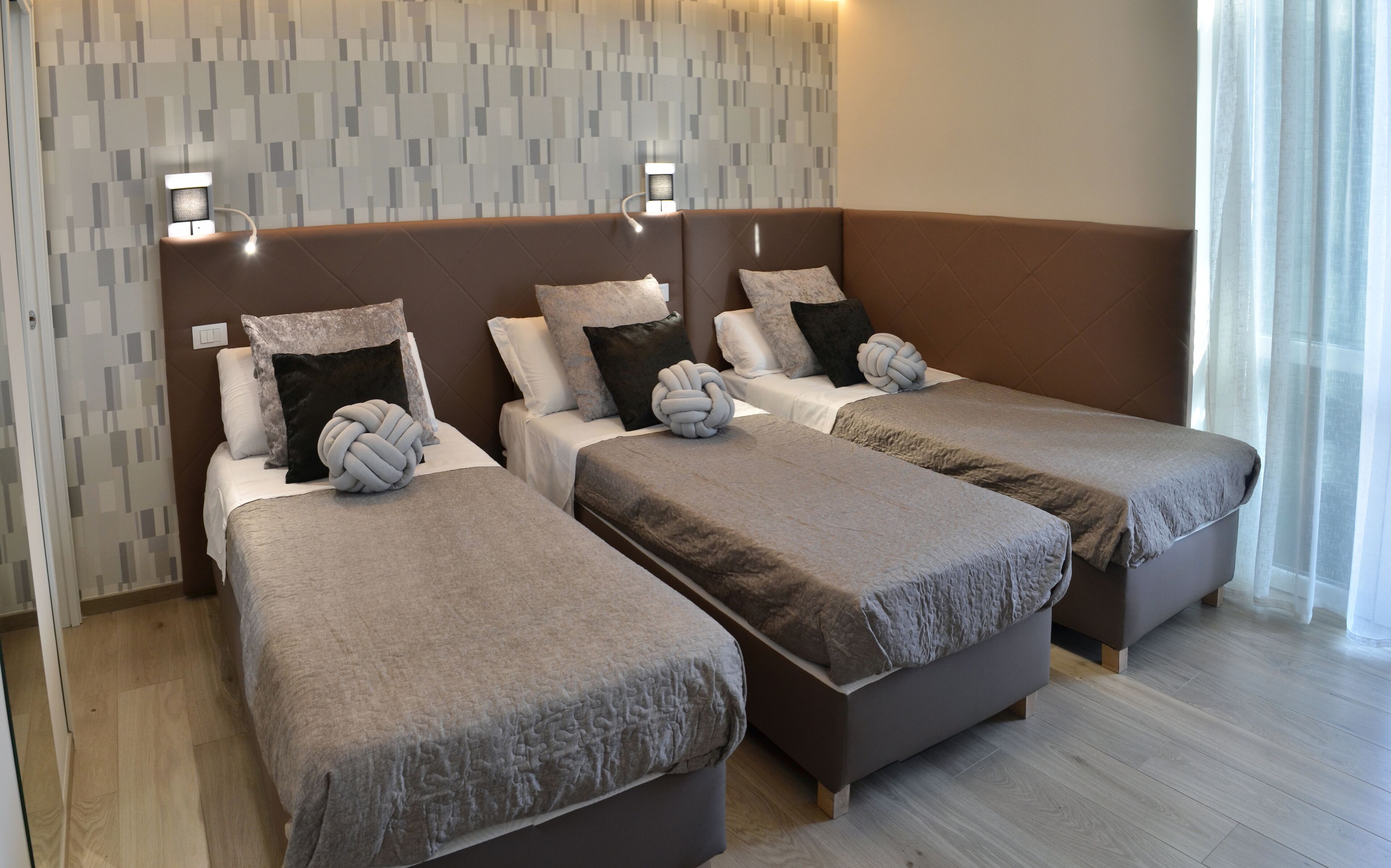 Habitación de hotel con dos camas individuales, cada una con una manta gris y un almohadón esférico. La habitación presenta un diseño moderno, con una pared estampada y un ventanal con cortinas de gasa.