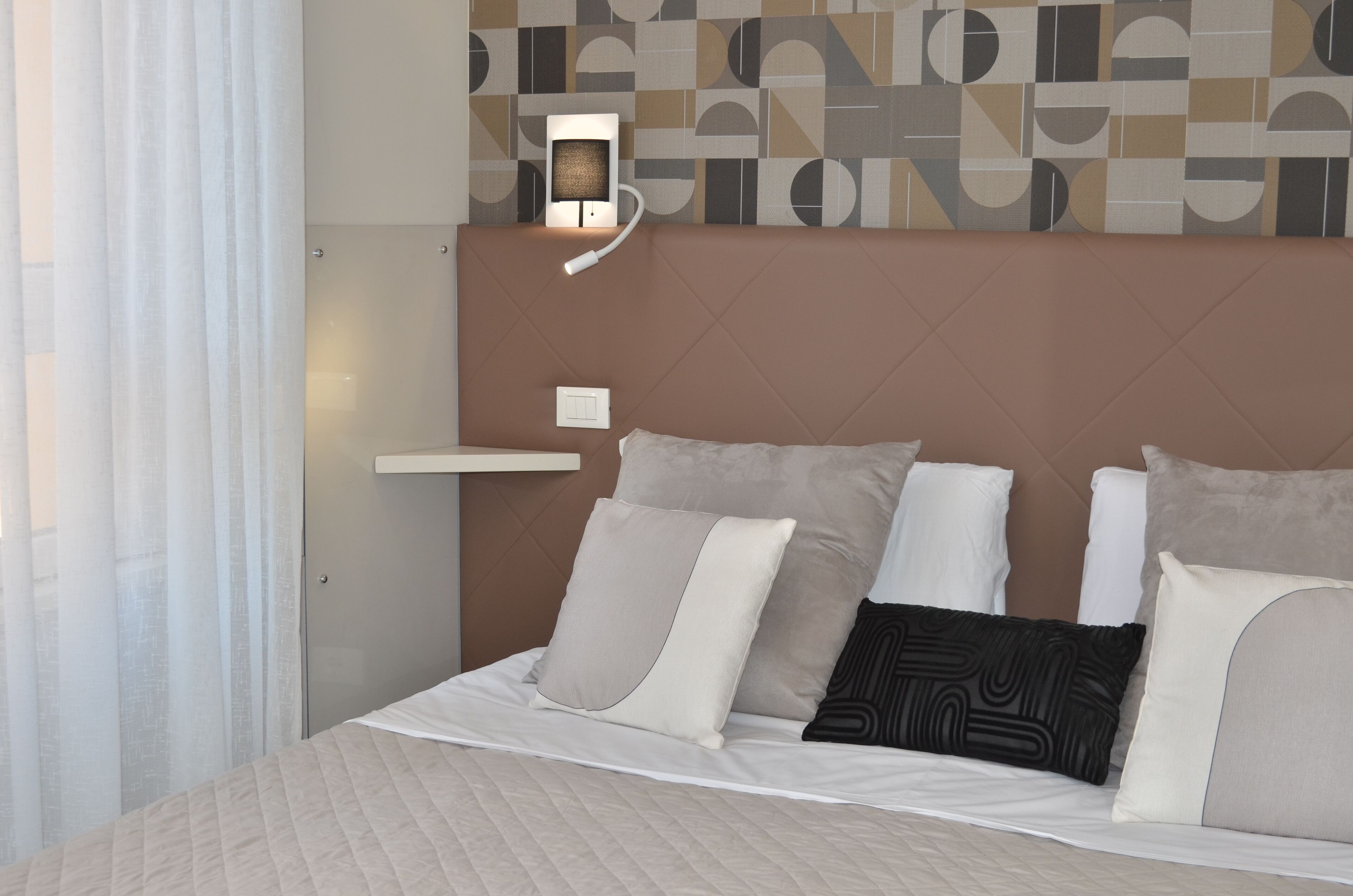 A hotel room with a bed, pillows, a bedside table, a wall-mounted light, and a patterned wall.