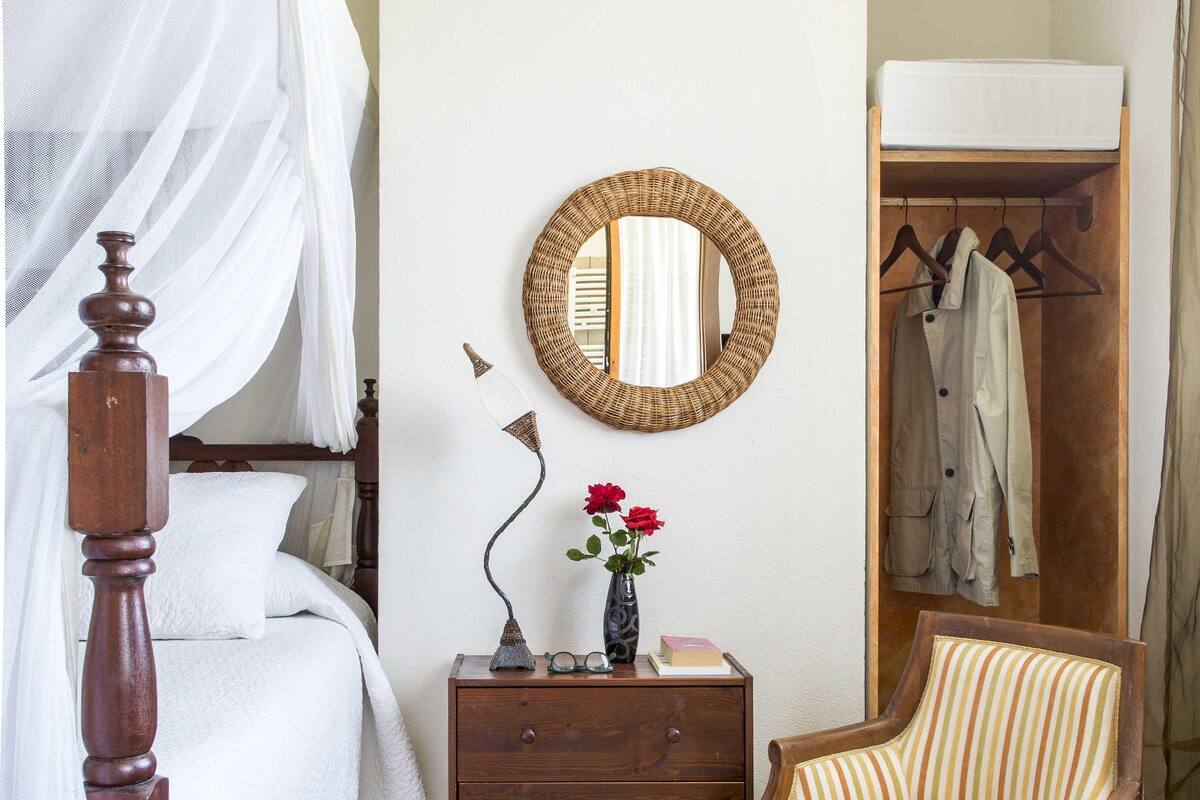 A bedroom with a four-poster bed, a wooden nightstand, a striped armchair, a mirror, and a wardrobe.