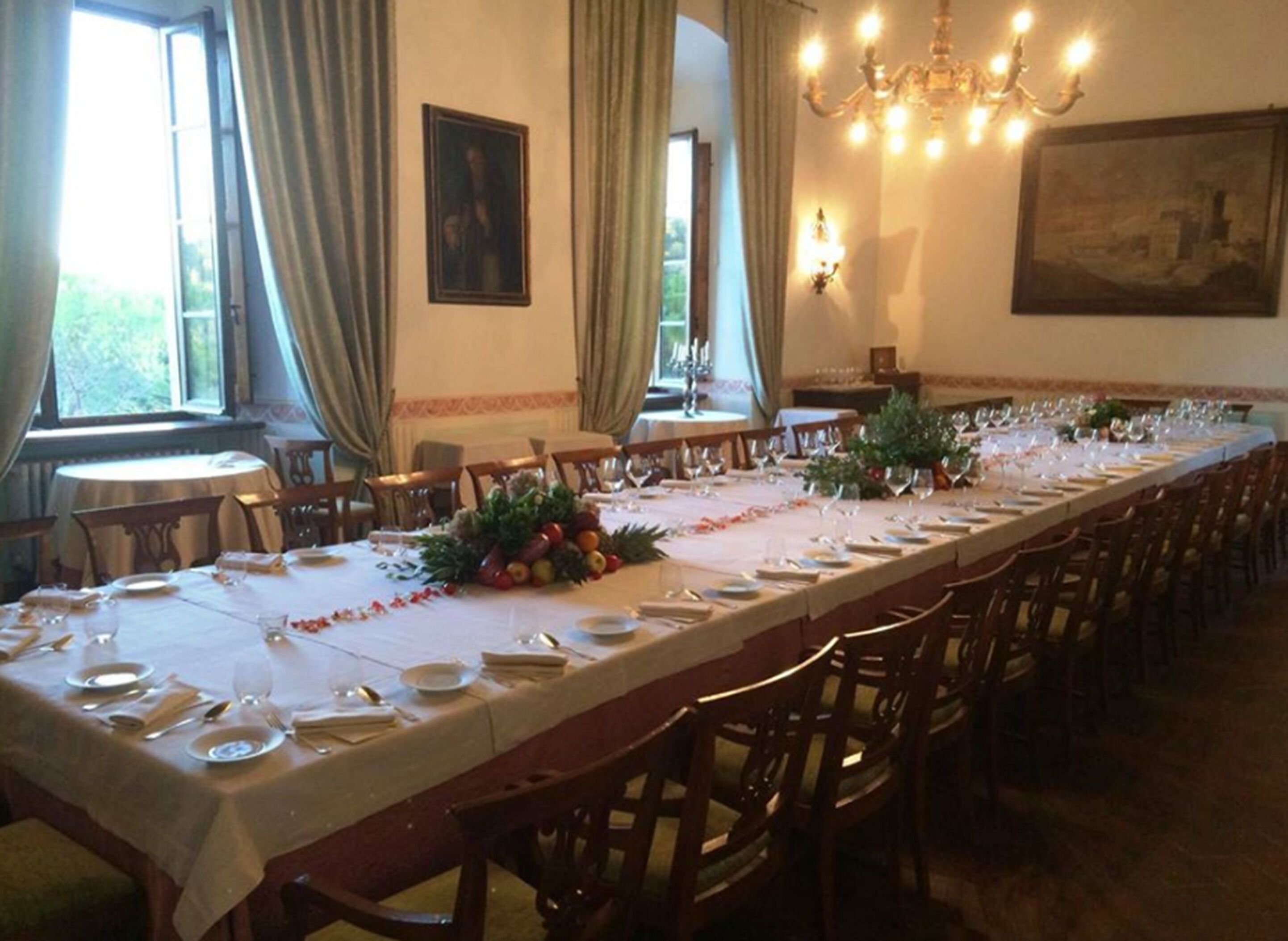 Une longue table à manger, dressée pour un repas, entourée de chaises, avec un lustre au-dessus et un tableau accroché au mur.