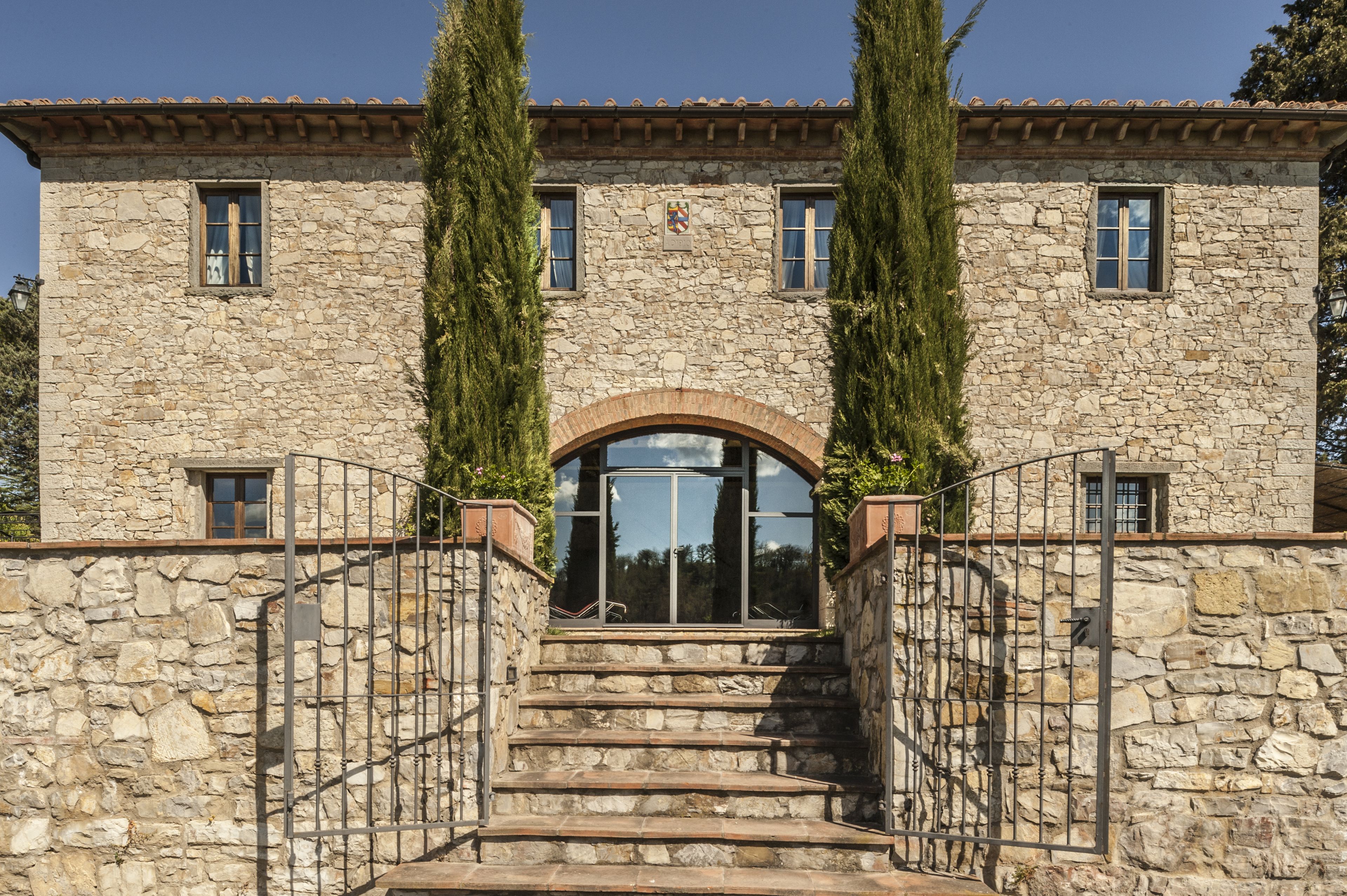Un edificio de piedra con una entrada central en arco, flanqueado por dos árboles altos y un portón metálico que conduce a la entrada.