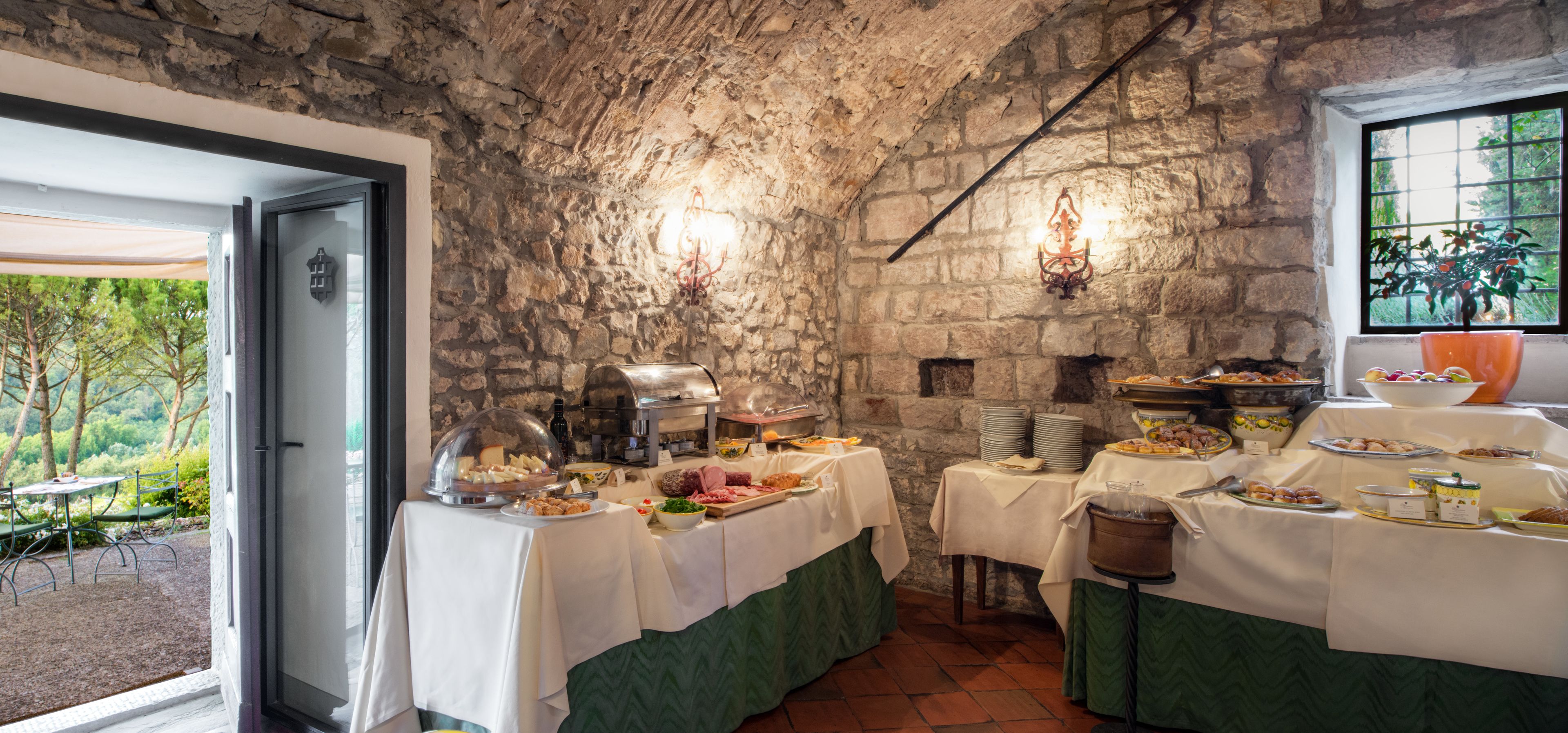 Un'area pranzo rustica con pareti in pietra, diversi cibi esposti sui tavoli e vista sul verde circostante.
