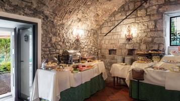 Un'area pranzo rustica con pareti in pietra, diversi cibi esposti sui tavoli e vista sul verde circostante.
