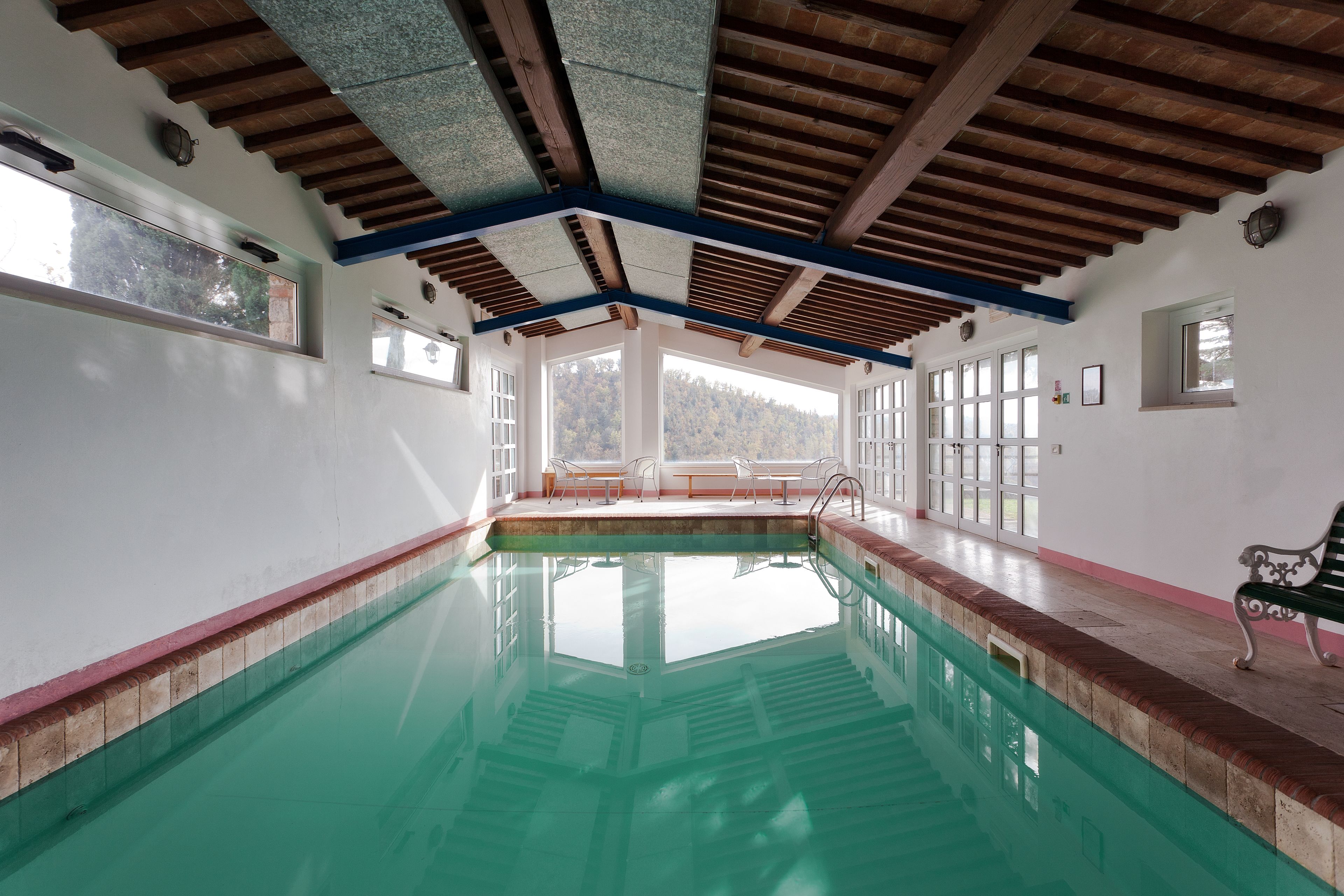 Una piscina interior con paredes de vidrio transparente, vigas de madera en el techo y un banco junto a la piscina.