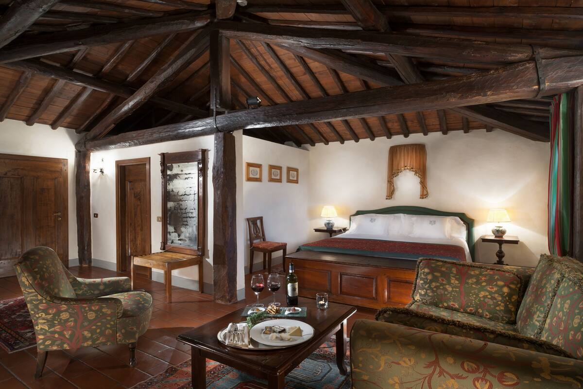 A traditional room with a wooden ceiling, a bed with a green bedspread, two upholstered chairs, a wooden table with a plate of food, and a wine bottle.