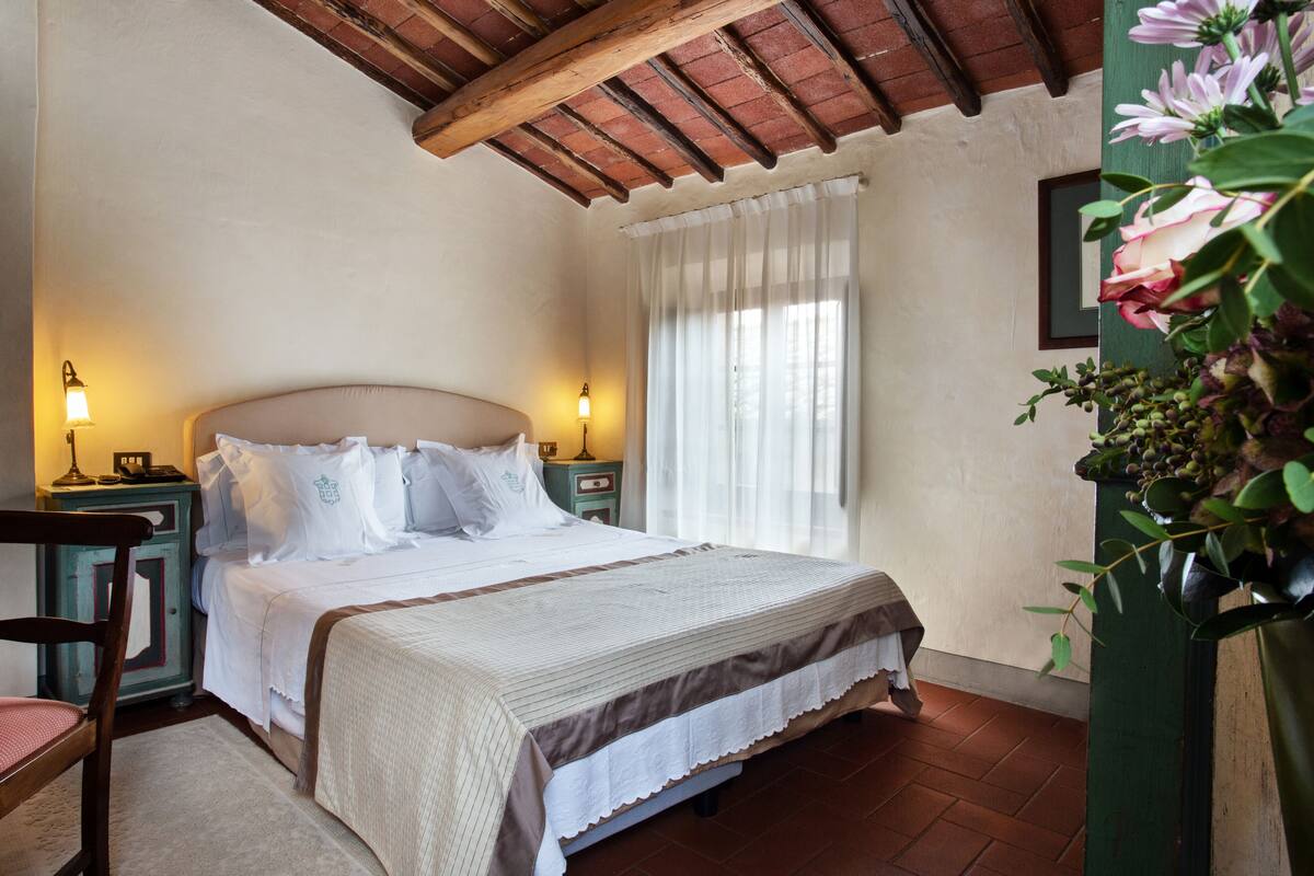 A bedroom with a bed, nightstands, a chair, a window with curtains, and a wall-mounted plant.