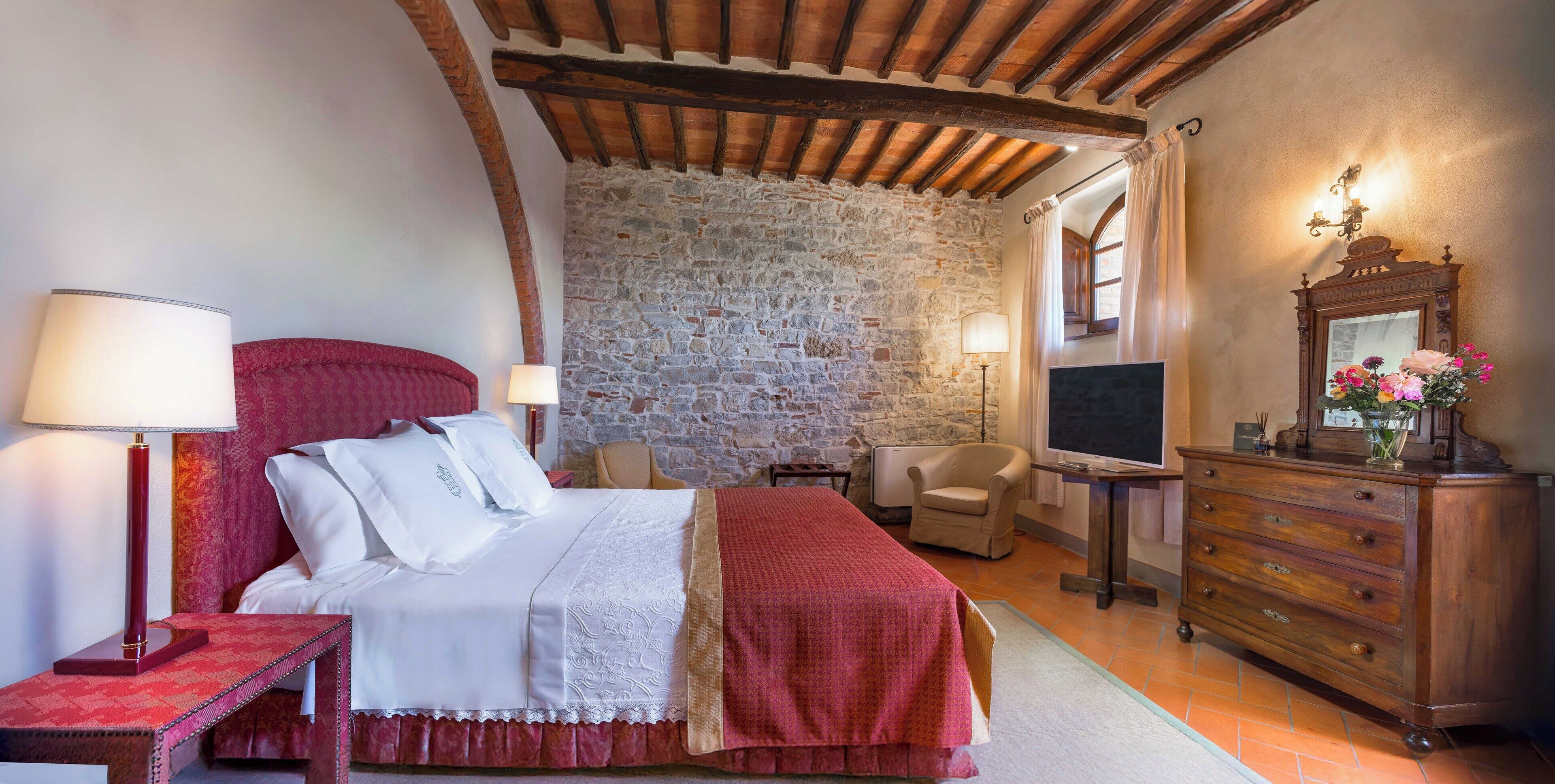 A bedroom with a stone wall, a bed with white and red bedding, a wooden dresser, a desk with a mirror, and a chair.