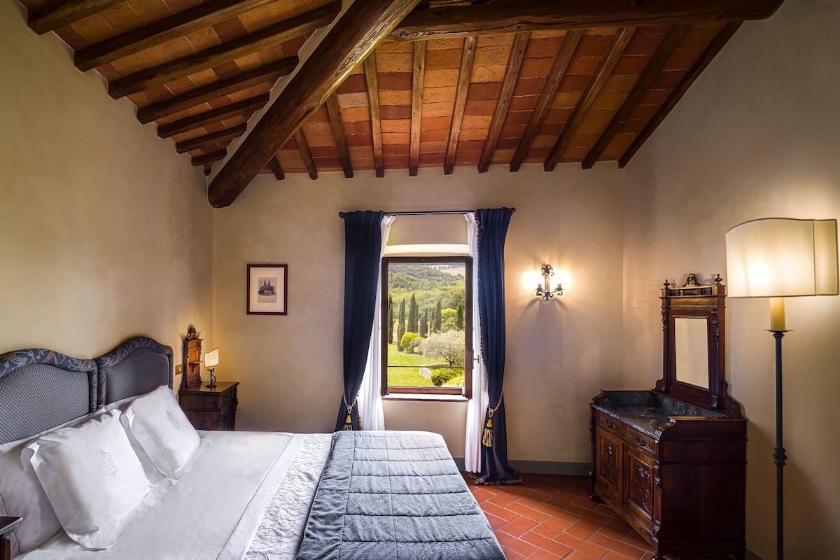 A bedroom with a wooden ceiling, a bed with white linens, a nightstand with a lamp, a dresser, and a view of greenery through a window.