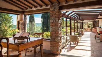 Une vaste terrasse dotée d’une table et de chaises en bois, de piliers en pierre et de grandes fenêtres offrant une vue sur le paysage environnant.