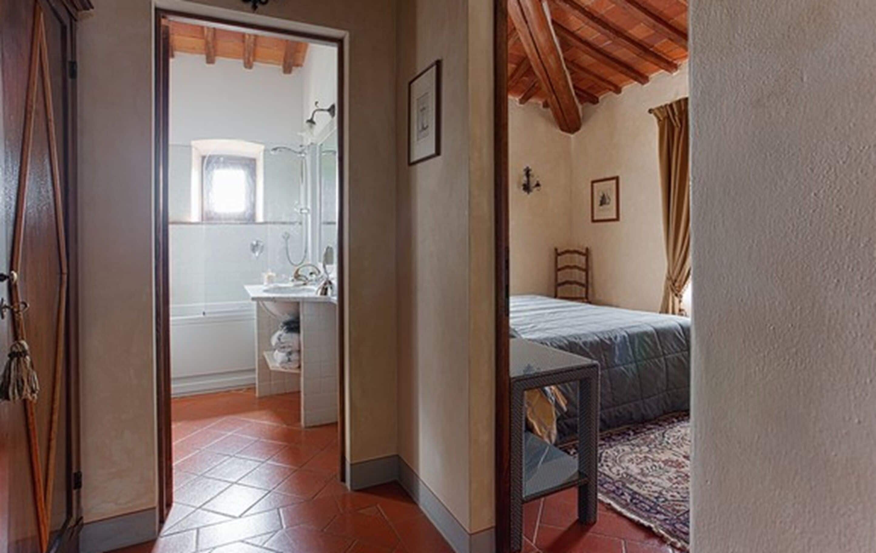 A bedroom with a bed, a small table, and a nightstand. There is a bathroom visible through an open door with a sink and mirror.