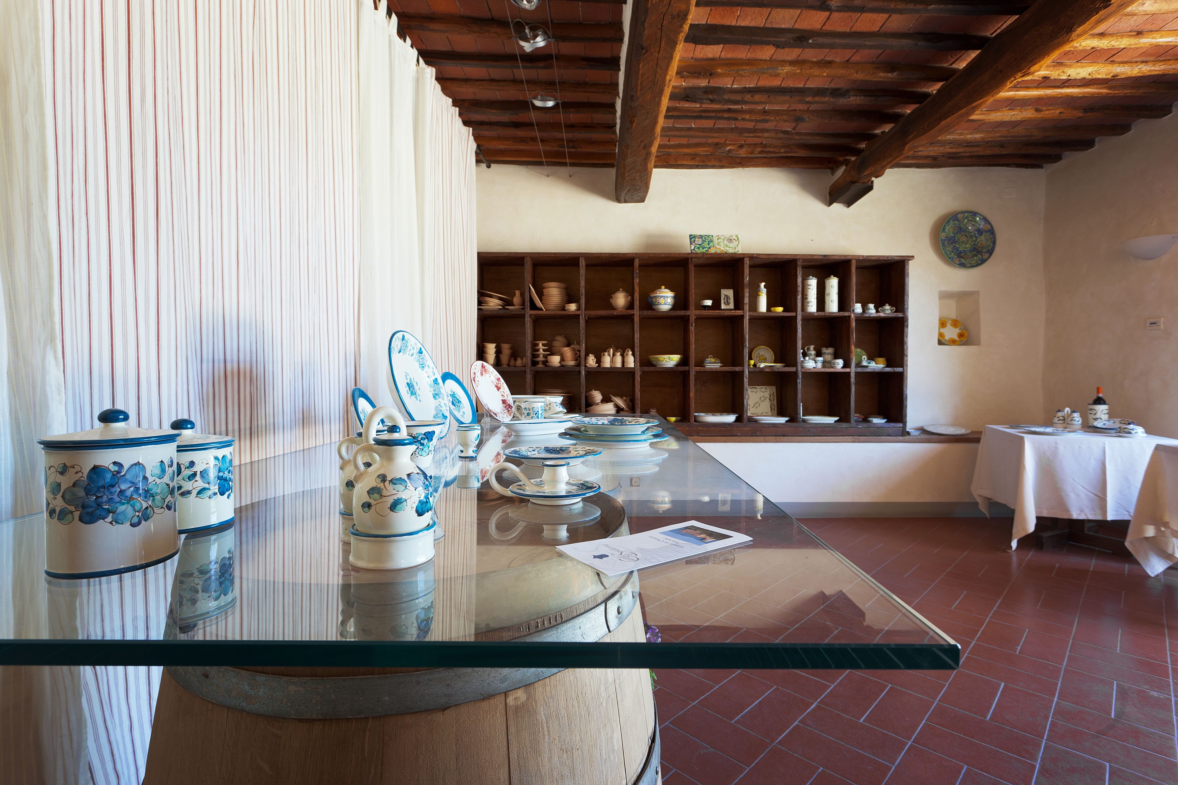 A room with a glass-top table displaying ceramic items, shelves with pottery, and a rustic wooden ceiling.