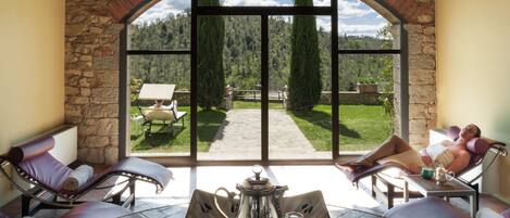 Ein Wintergarten mit Steinbögen, zwei Liegestühlen, einem Tisch mit Teekanne und Gläsern sowie Blick auf einen Garten und einen Weg.
