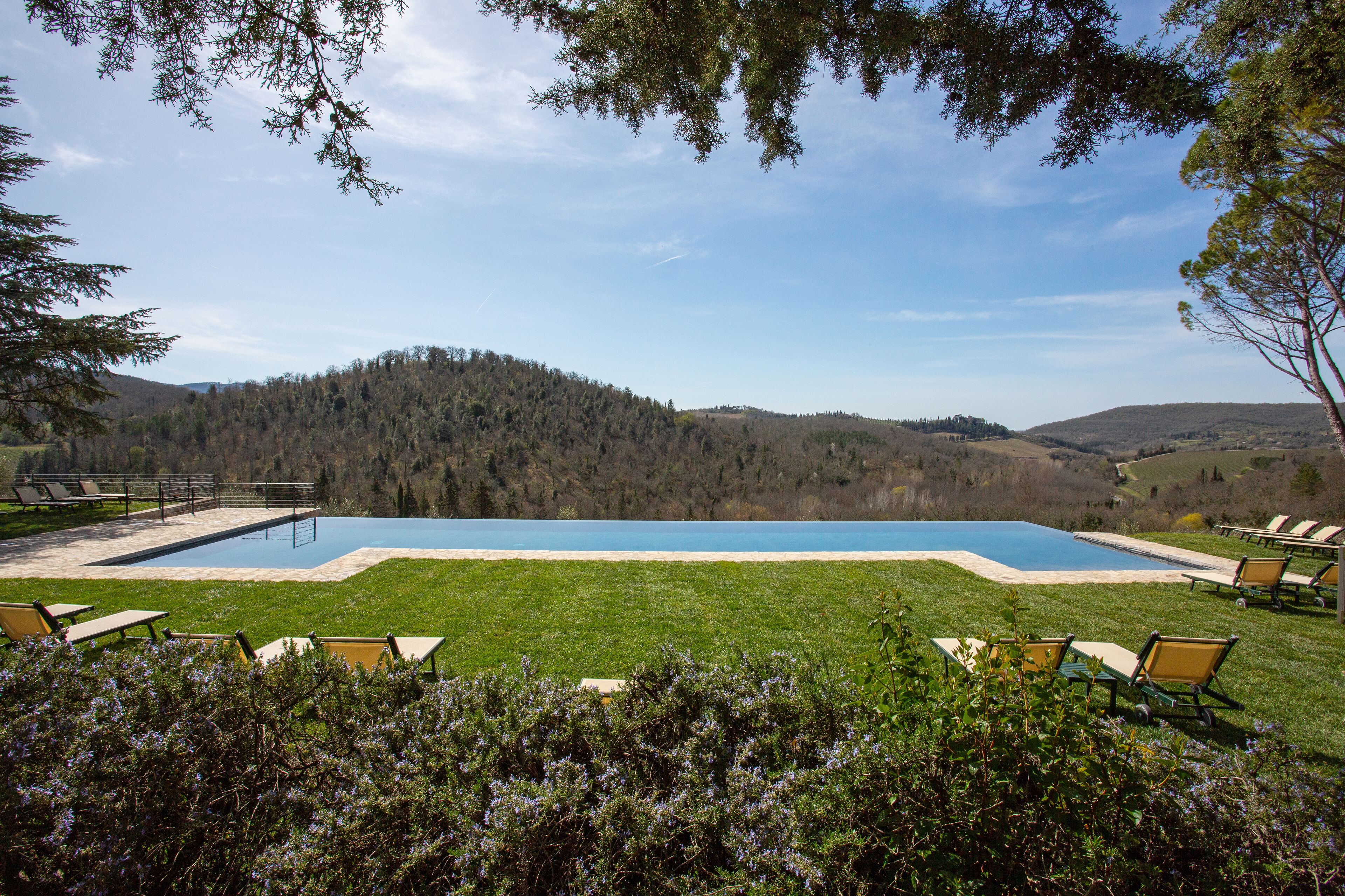 Una piscina con vistas a una colina boscosa, rodeada de sillas de jardín y una terraza.