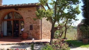 A stone building with a large arched doorway, a person standing inside, and a garden with flowers and a tree.