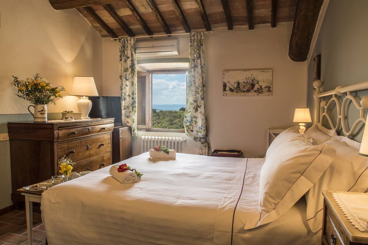 Un dormitorio con cama, cómoda, mesita de noche, lámpara, ventana con cortinas y vistas al exterior.