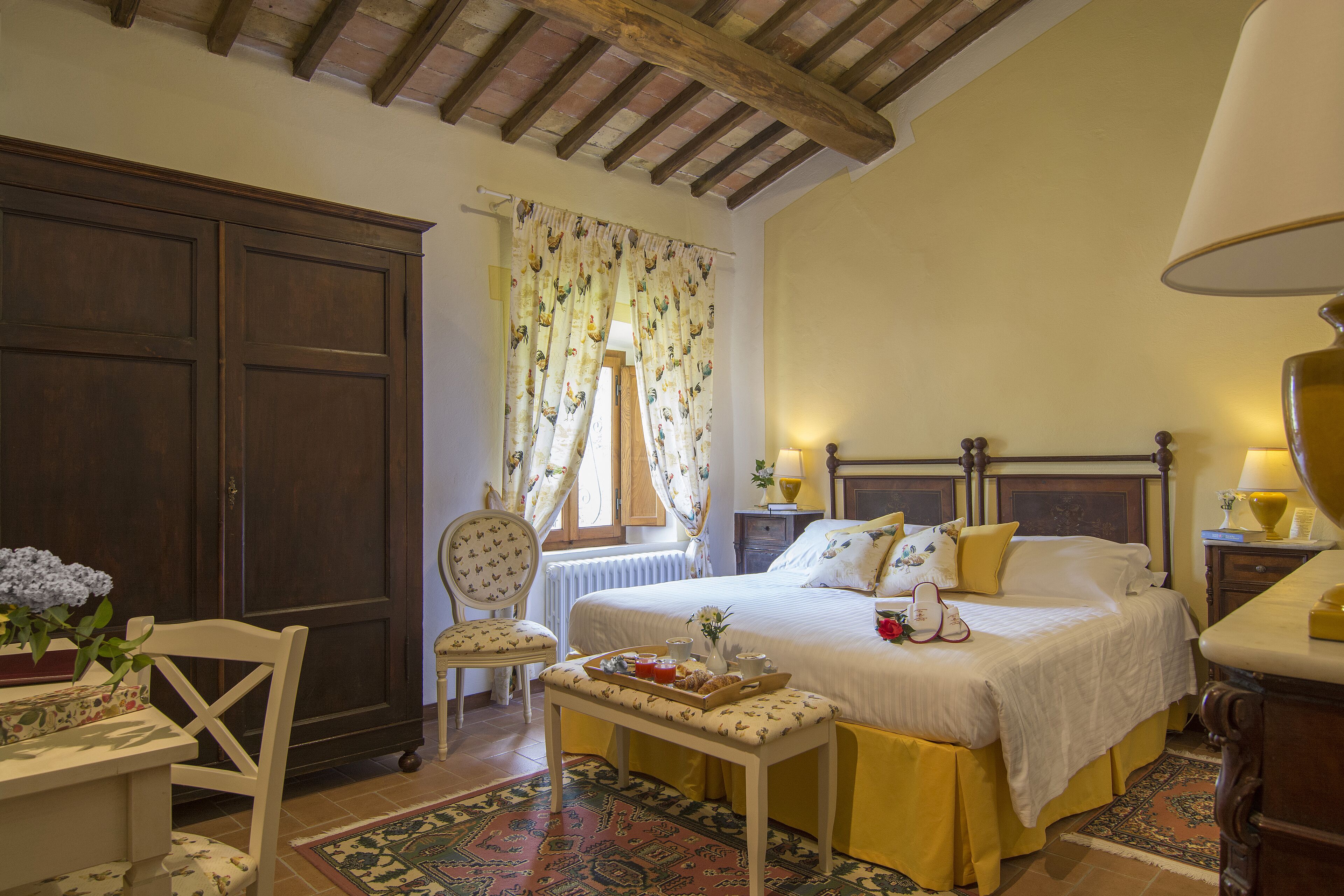 A bedroom with a large bed, a wooden wardrobe, a chair, a table with a tray, and a window with curtains.