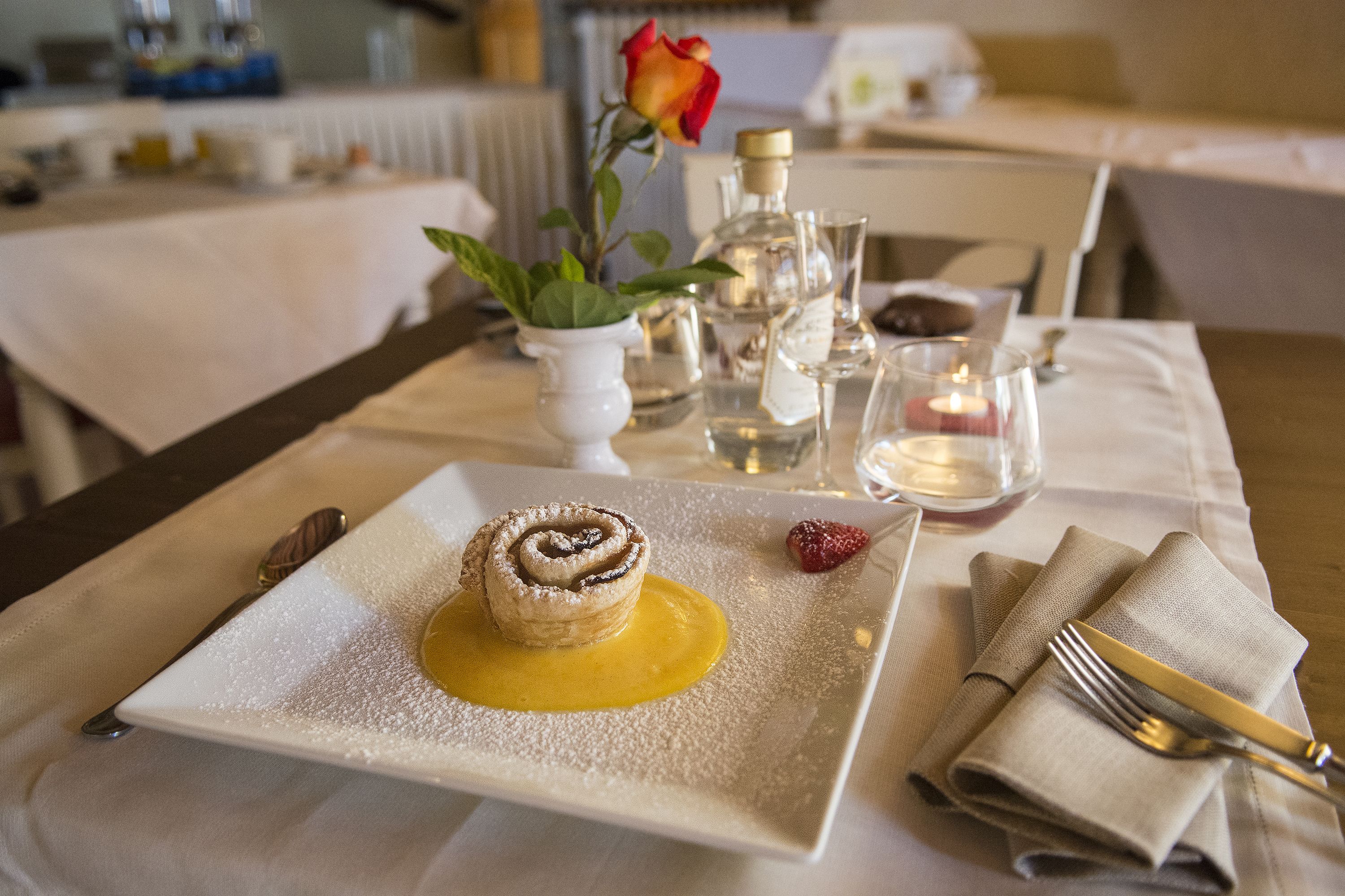 Ein Dessert auf einem quadratischen Teller, im Hintergrund eine Kerze und eine Rose.