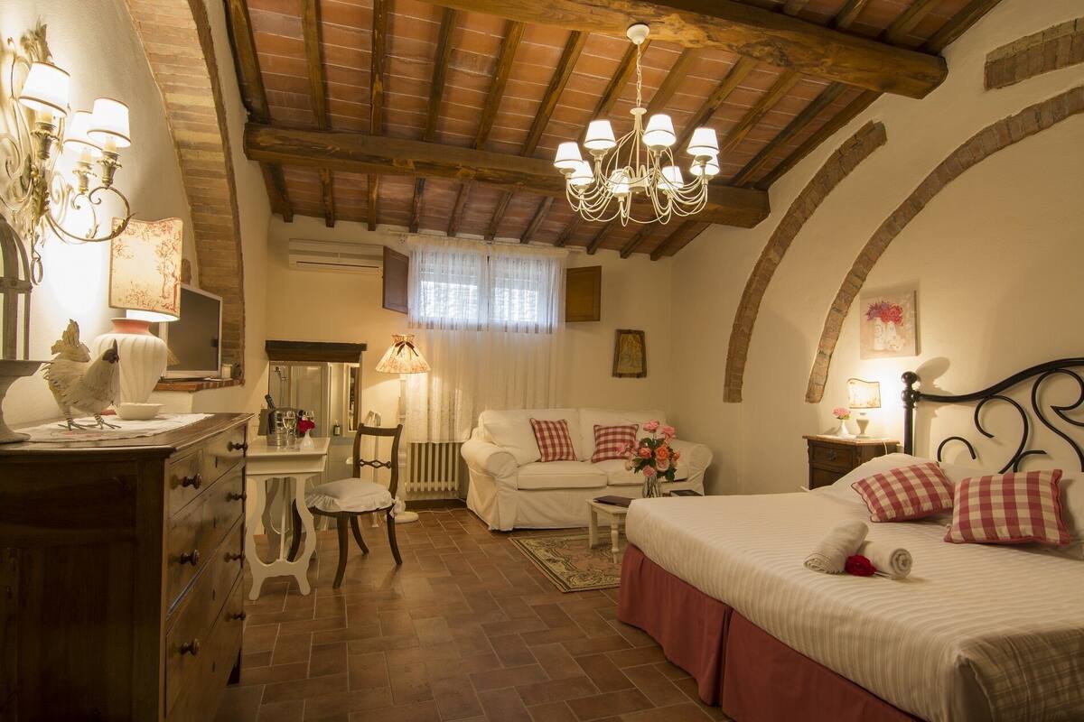 Un dormitorio acogedor con techo de madera, cama blanca con almohadas rosadas, sofá blanco, cómoda de madera y candelabro.