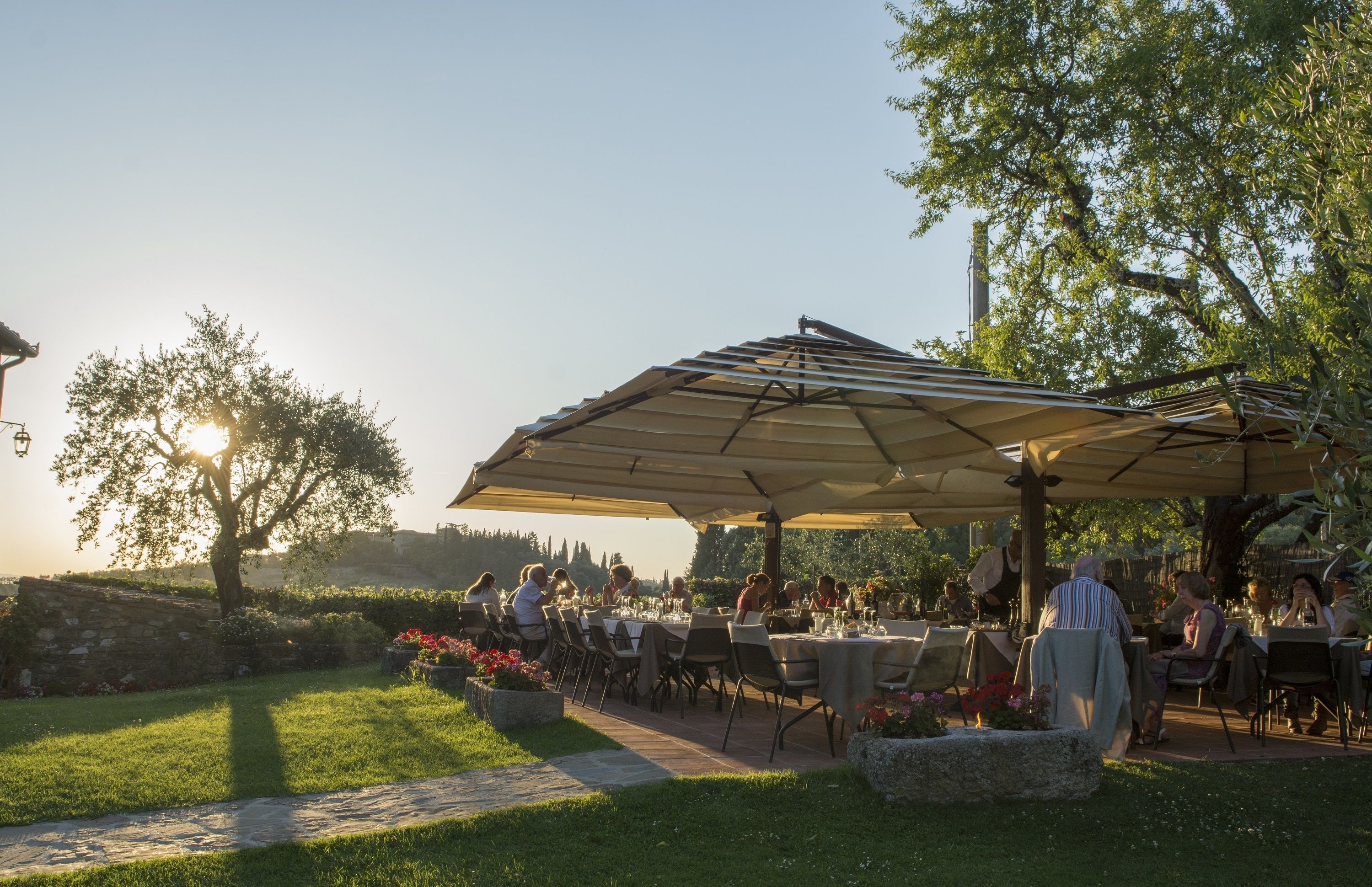 Un área de comedor al aire libre con mesas y sillas bajo un gran paraguas, rodeada de follaje y un muro de piedra.