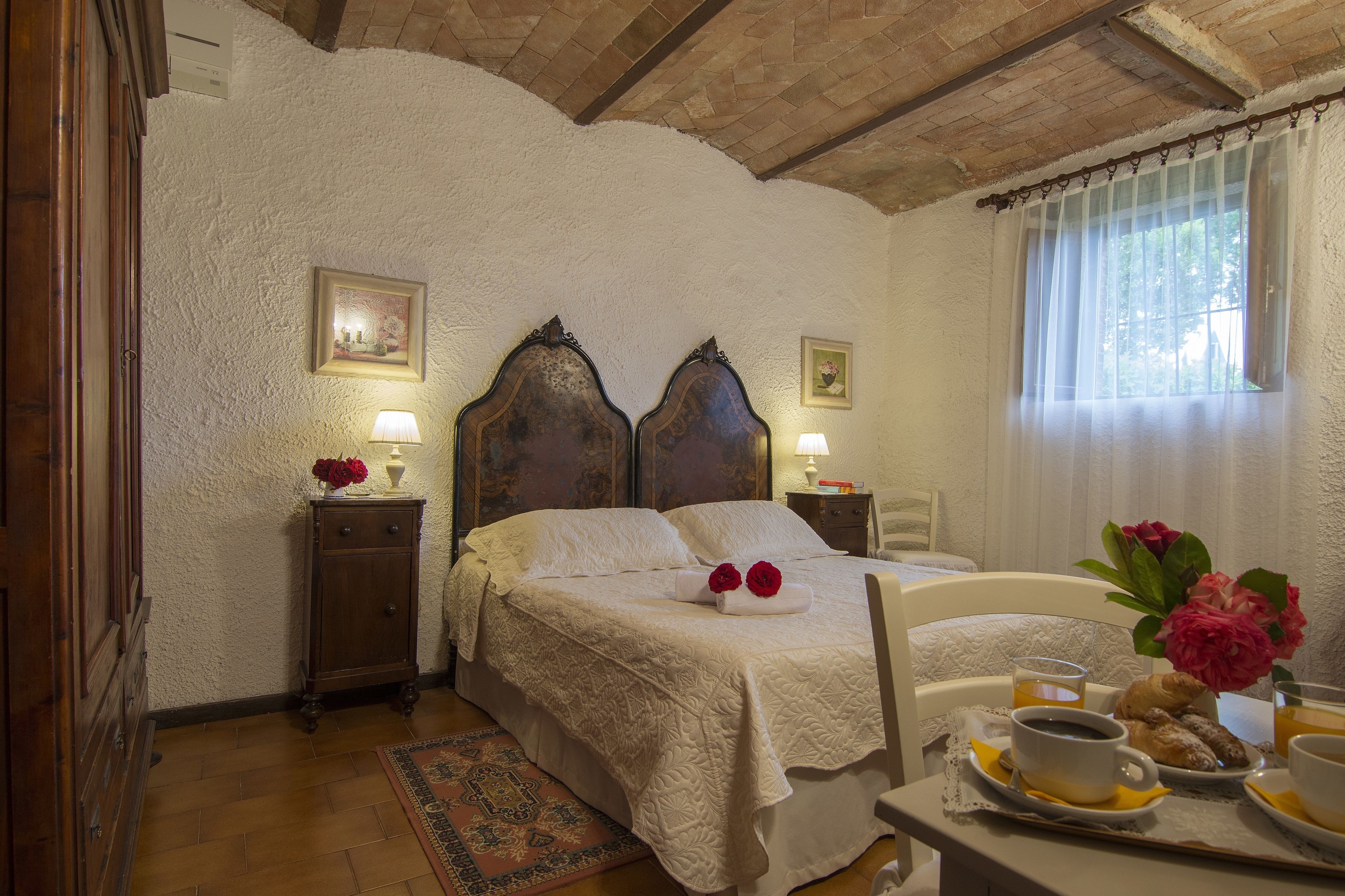 A bedroom with a large bed, bedside tables, a nightstand, a window with curtains, and a breakfast tray with coffee, pastries, and flowers.