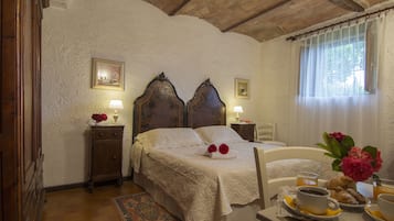 A bedroom with a large bed, bedside tables, a nightstand, a window with curtains, and a breakfast tray with coffee, pastries, and flowers.