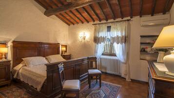Un dormitorio con cama de madera, dos sillas, una mesita de noche, una lámpara, un radiador y una ventana con cortinas.