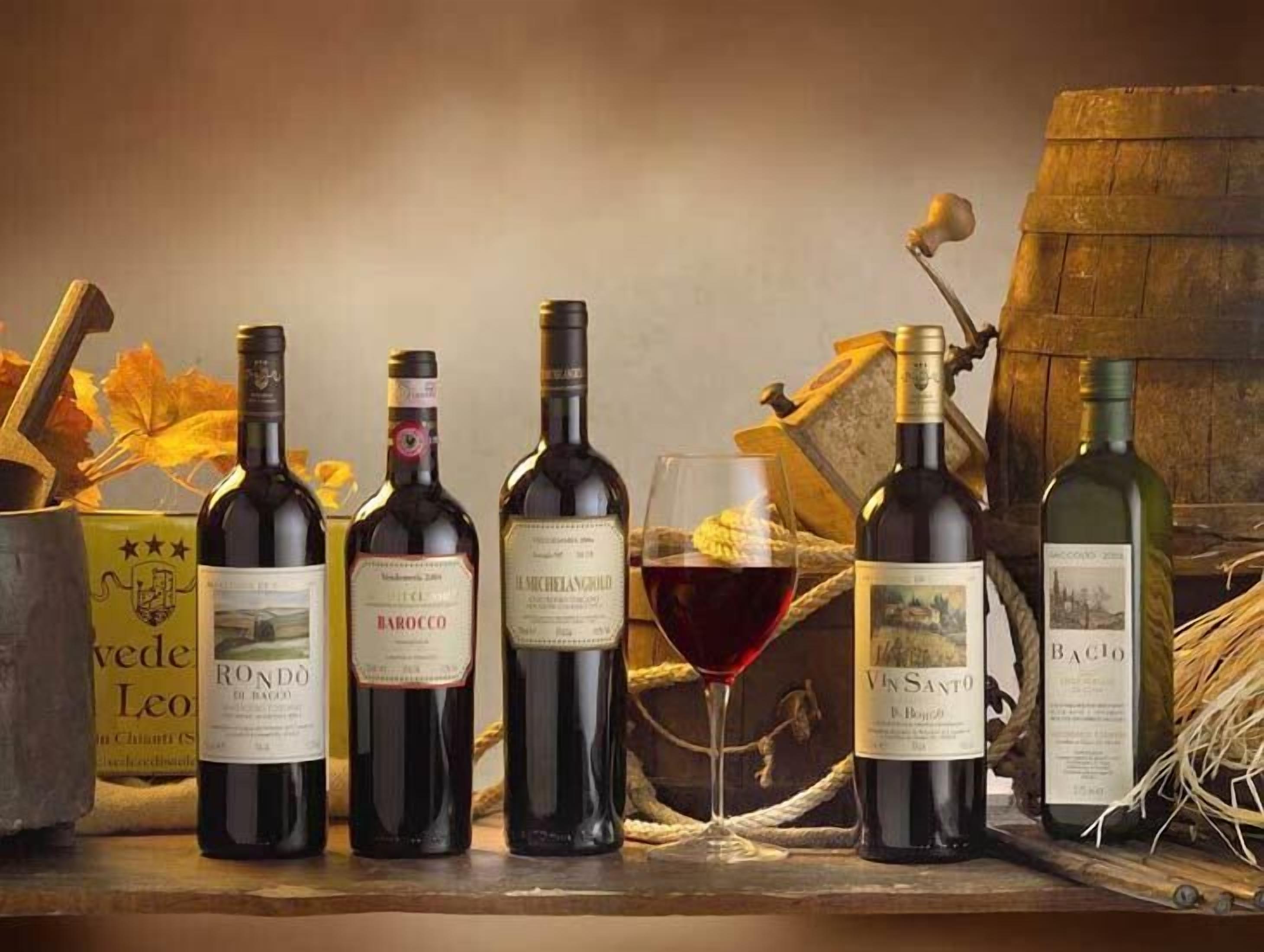 A selection of wine bottles and a glass of red wine on a wooden surface with autumn leaves and a barrel in the background.