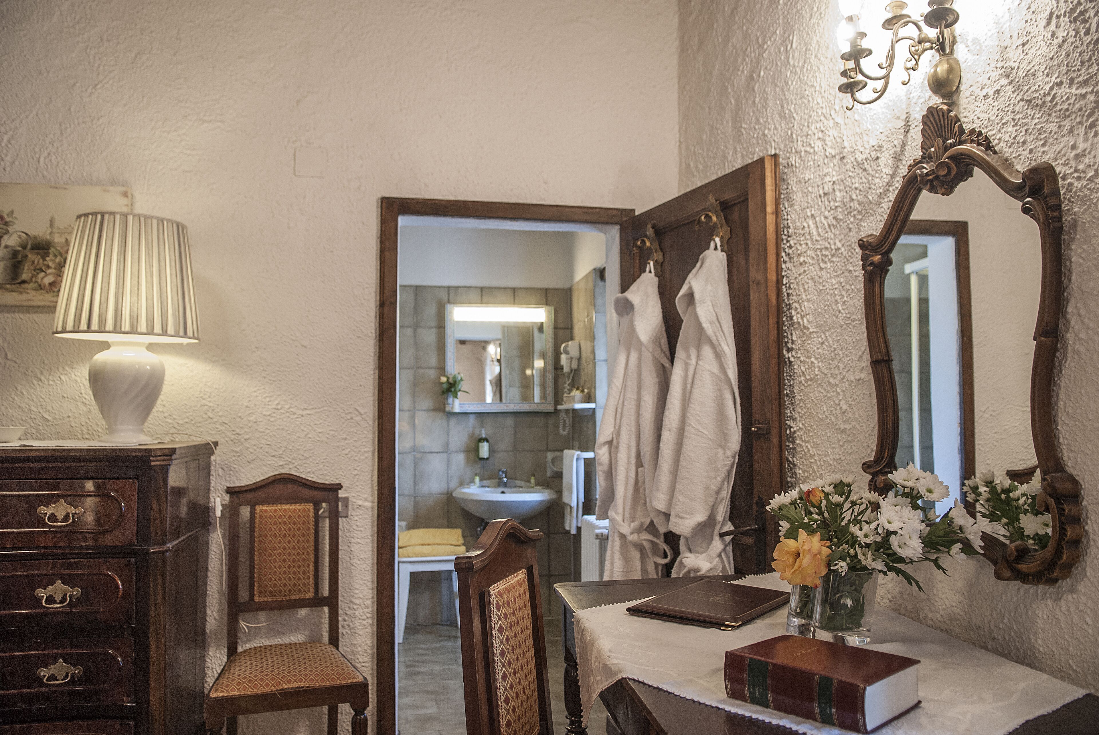 Une salle de bain avec une chaise en bois, une table sur laquelle se trouvent un livre et un vase, et un miroir doté d’un cadre décoratif.