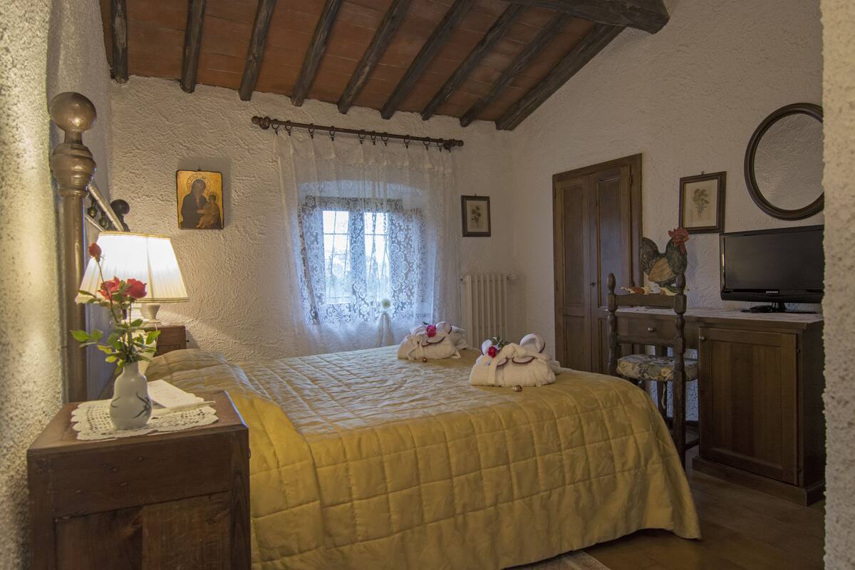 A bedroom with a bed, a nightstand, a lamp, a window with lace curtains, a radiator, a wooden door, a television, and a wall-mounted picture.