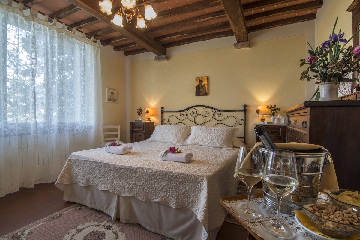 A neatly made bed with a decorative headboard, a bedside table with a lamp, and a tray with wine glasses and snacks.