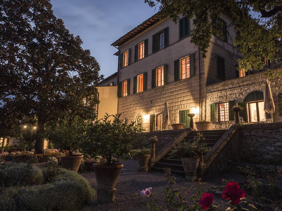 A historic building with a stone facade, multiple windows with shutters, and a well-maintained garden with potted plants and flowers.