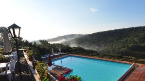 Une piscine d’un bleu éclatant, bordée de chaises longues et d’ombrelles, entourée d’une végétation luxuriante et d’une colline boisée.