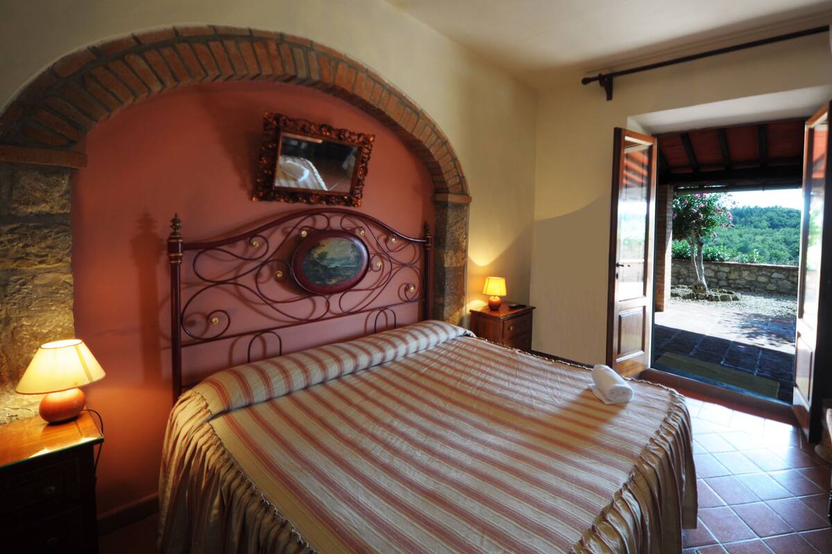Un dormitorio con cama, mesita de noche, lámpara y vista al exterior a través de una puerta abierta.