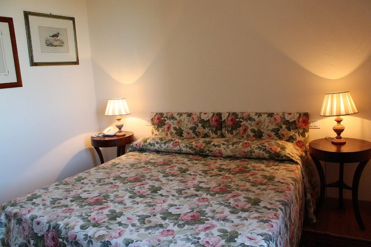 Une chambre à coucher avec une couvre-lit à motifs floraux, deux lampes de chevet, un téléphone sur la table de chevet et des œuvres d’art encadrées accrochées au mur.