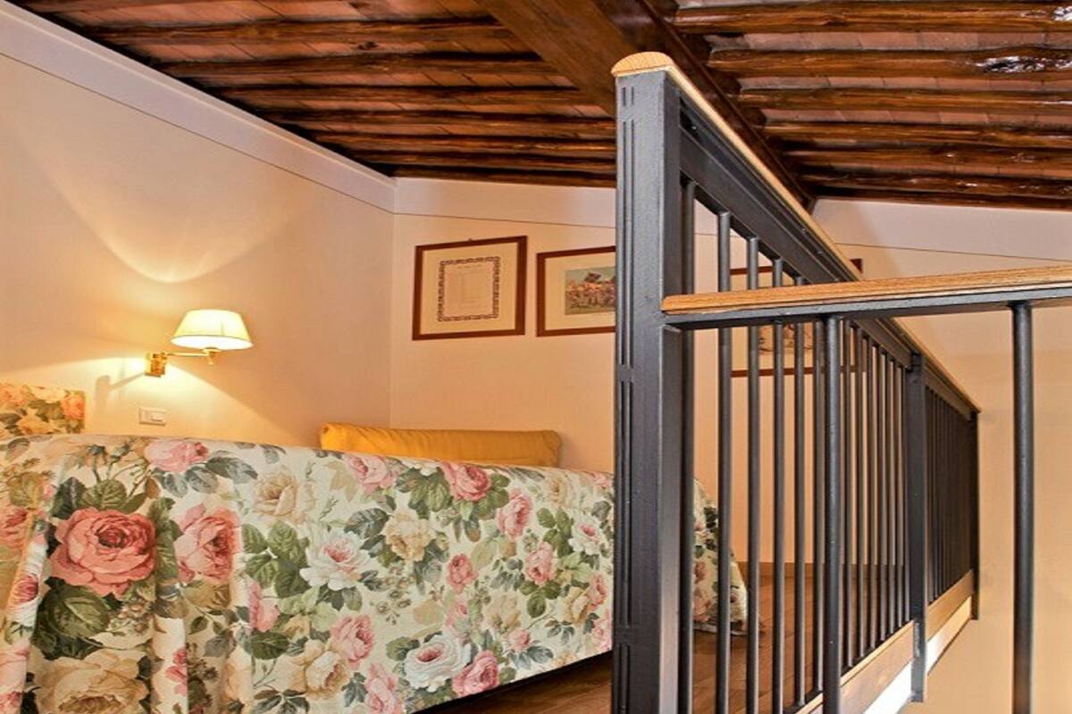 Une chambre avec un plafond en bois, un lit recouvert d’une literie à motifs floraux, une table de chevet et une lampe fixée au mur.
