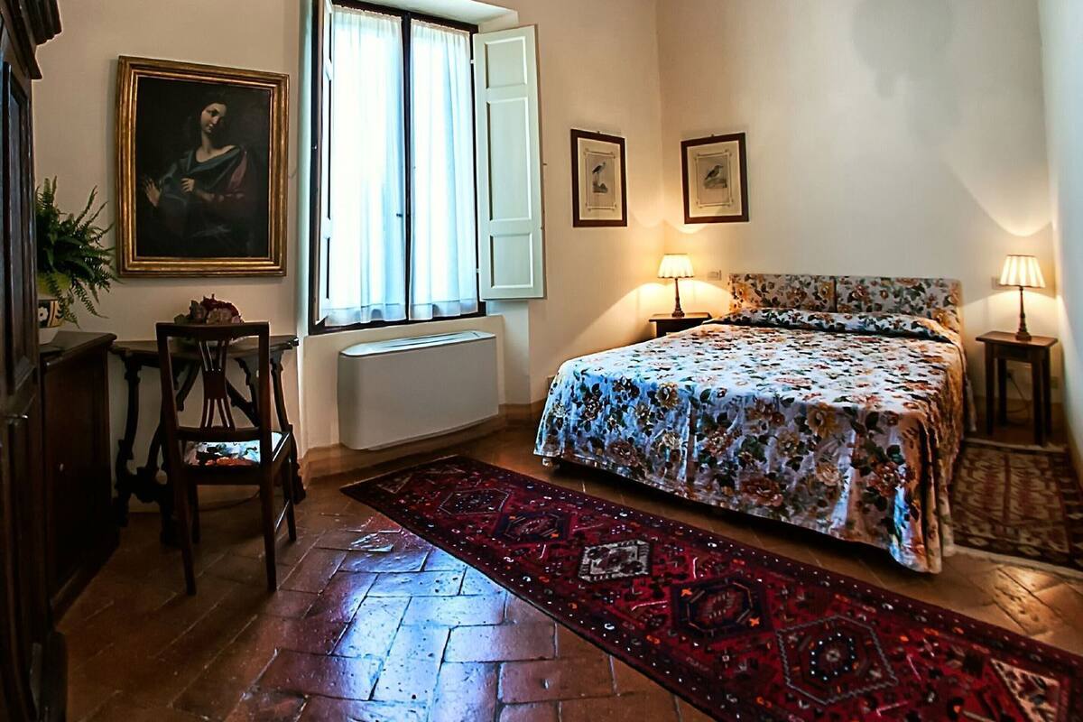 Une chambre avec un lit, deux lampes de chevet, une fenêtre avec des rideaux, un radiateur, un tableau encadré et un tapis à motifs.