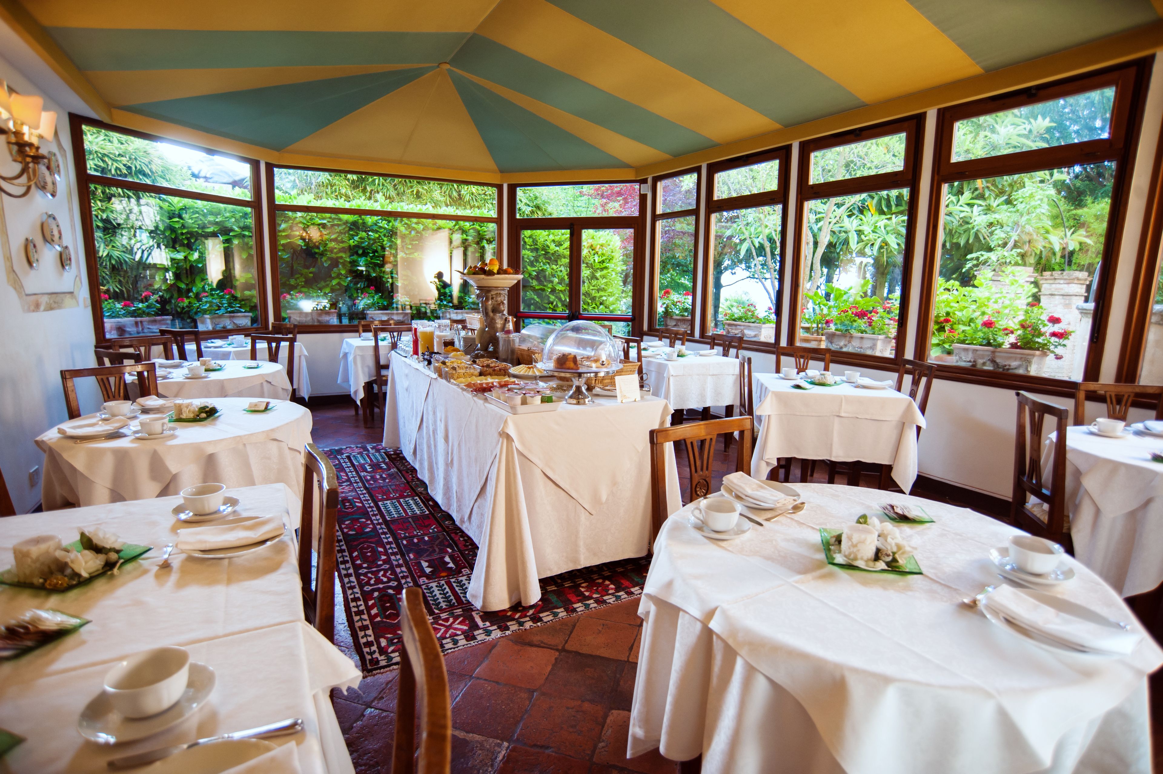 Ein Essbereich mit runden Tischen, gedeckt für ein Frühstück, große Fenster mit Gartenblick und eine zentrale Buffetstation.
