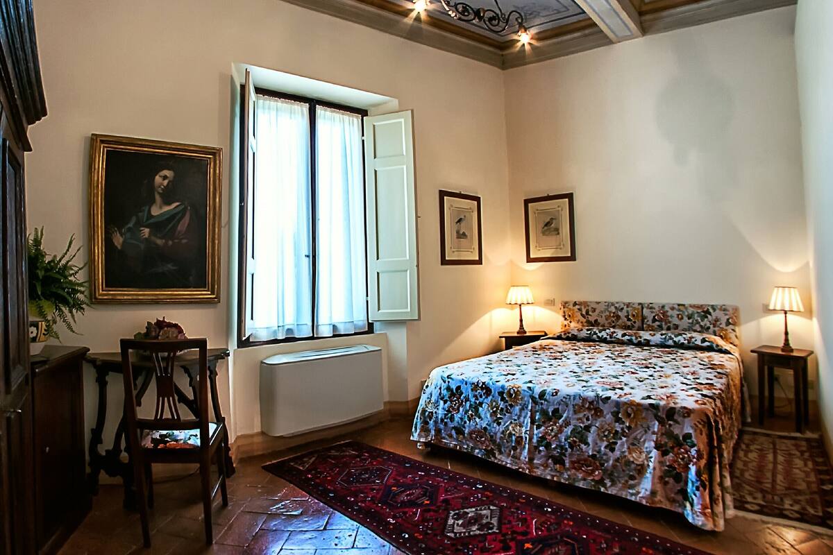Une chambre à coucher avec un lit, deux lampes de chevet, une fenêtre avec des rideaux, un radiateur, une chaise, un tableau encadré et un tapis à motifs.