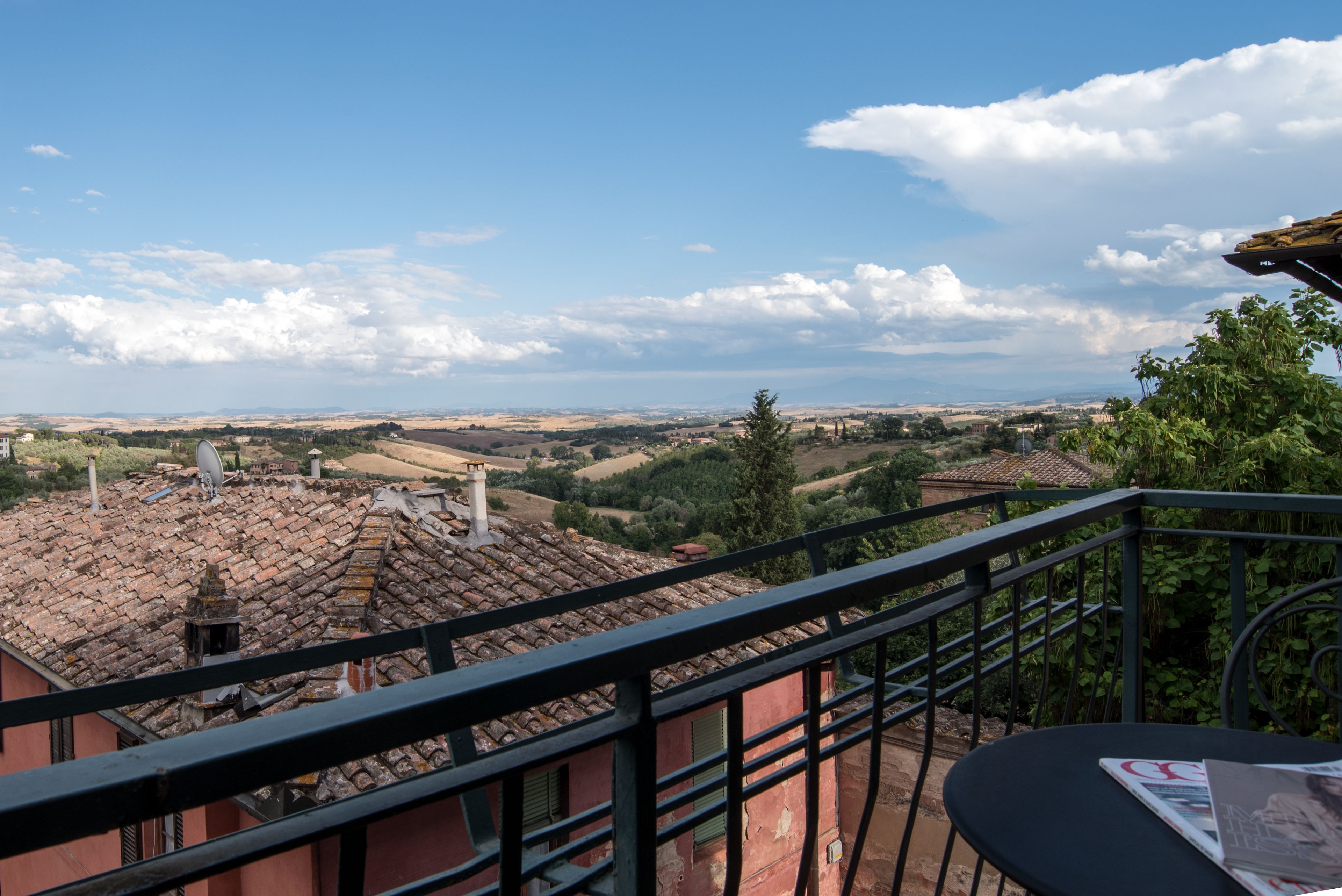 A balcony with a view of terracotta-roofed buildings, a table with a magazine, and a clear sky.