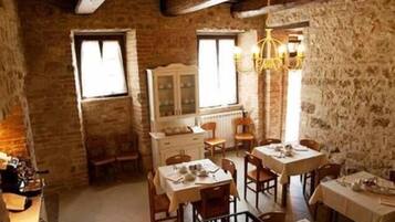 A rustic dining area with stone walls, wooden beams, and a chandelier. Tables are set with plates and cutlery.