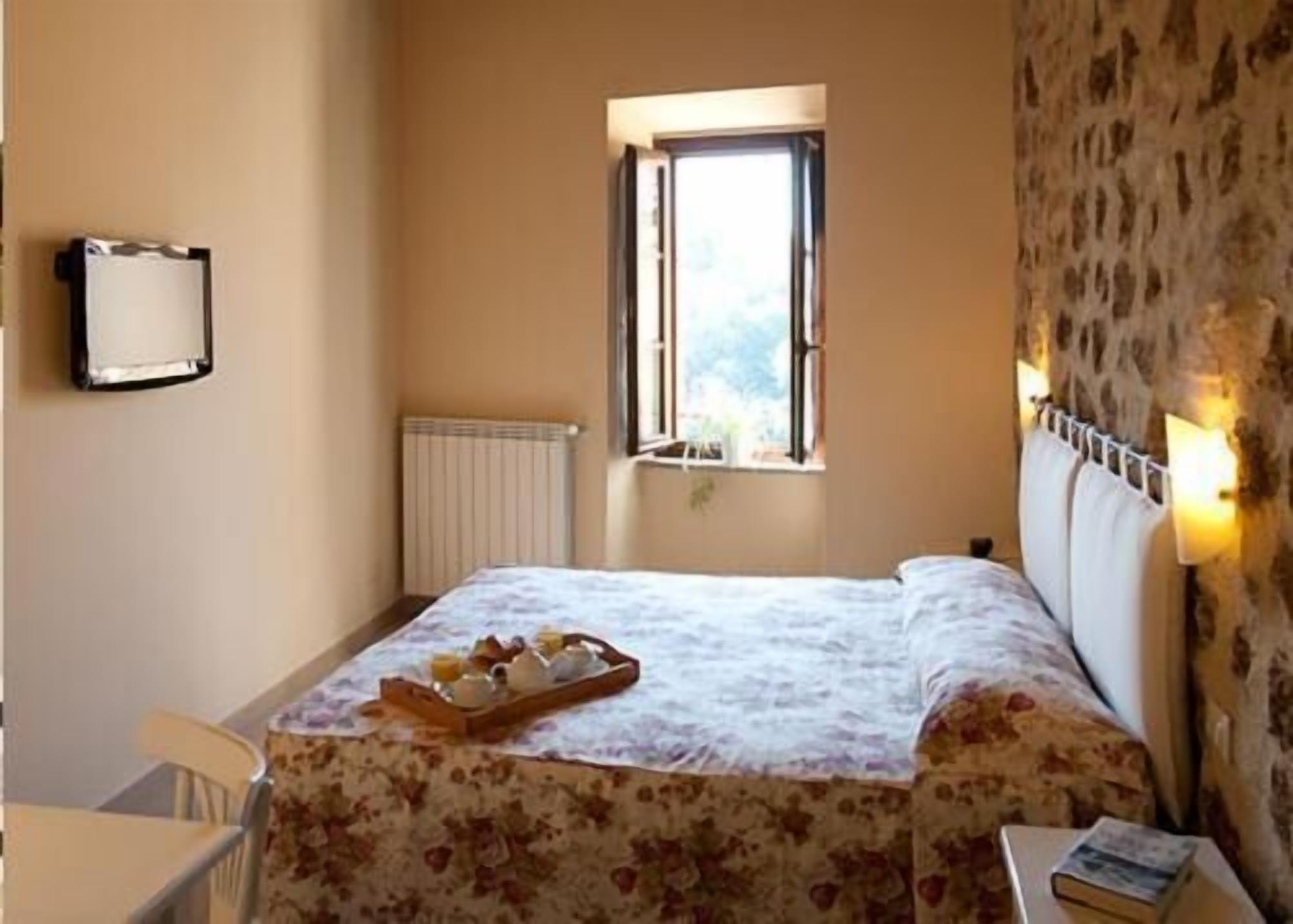 Un dormitorio con cama, ventana, radiador, mesita de noche y estante fijado a la pared.